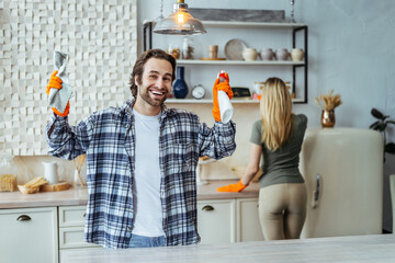 Happy young european wife and husband in rubber gloves raise hands with cleaning supplies, clean...