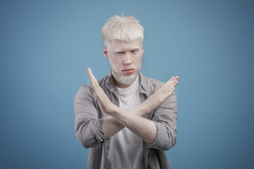 Serious young albino guy saying no, looking at camera and showing hands crossed over chest, blue...