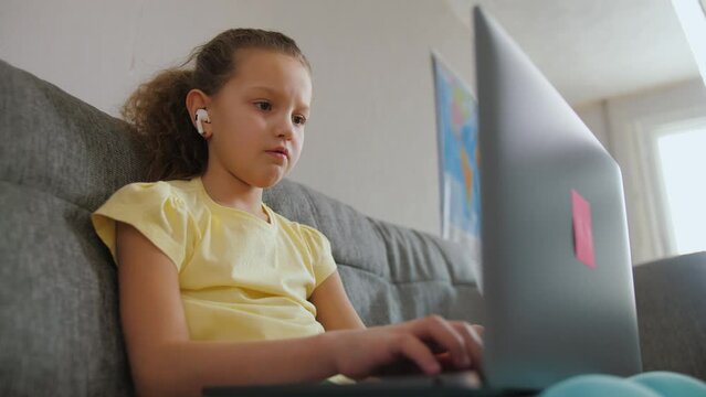 Cute primary school child girl studying with laptop at home, sit on sofa and doing homework. Little kid with headphones listening audio lesson, typing on keyboard, elementary education, close up