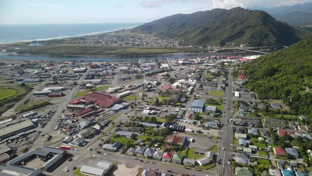 Greymouth town, aerial pull back reveal of small city on West Coast, commercial and housing area, traffic. Mountain and seaside background.