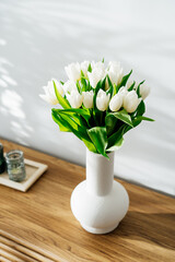 Spring bouquet of white tulip flowers in vase stand on wooden table near light grey wall with highlights and shadows. Gift for holiday, birthday, 8 March, Mother's Day. Vertical card. Selective focus.