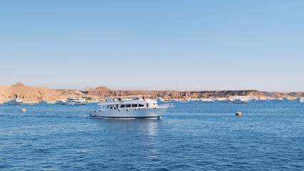 Obraz premium Large white yacht sailing across blue sea on sunny day, against marina backdrop with anchored yachts. Luxury motor yacht side view emerging from ship dock. Egypt Red Sea Cruise. Holiday yacht vacation