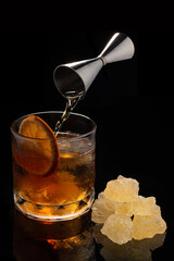 Alcoholic cocktail Old fashioned cocktail with orange slice, spilling bourbon from the double jigger and lump sugar on black background. Copyspace