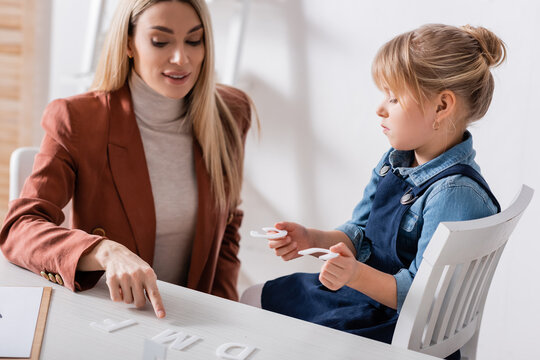 Speech therapist pointing at letter on table near kid in consulting room.