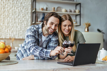 Happy young european man with stubble and lady clink glasses with wine and look at laptop in modern kitchen