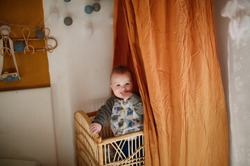 Cute baby in a wicker crib and under a terracotta canopy. Baby is standing in bed