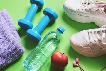 Purple towel, blue dumbbells, water bottle, apple, sneakers and headphones on a green background. Gym, healthy lifestyle flat lay photography. Fitness accessories and proper nutrition