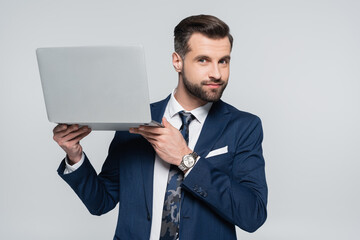 positive businessman looking at camera while holding laptop isolated on grey.