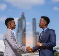 businessmen hand shake together, building in city with light at background. black man in grey suit and executive man with glasses in blue suit