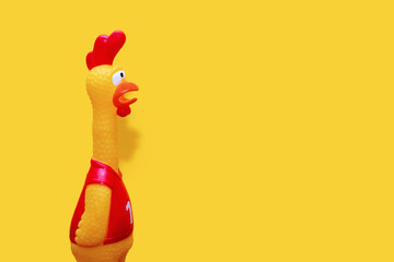 Rubber toy in the form of a rooster on a yellow background. The funny toy rooster has a surprised...