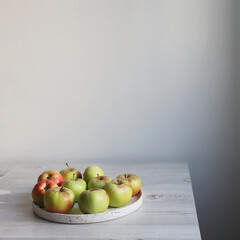 The green and red apples in a white faience bowl on the edge of a beige kitchen table. Scandinavian style. Minimalism. Place for text.