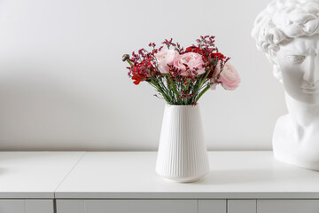Bouquet of red and pink Persian buttercups and plaster head of a Greek hero on a white table. Scandinavian style. Place for text. Copy space