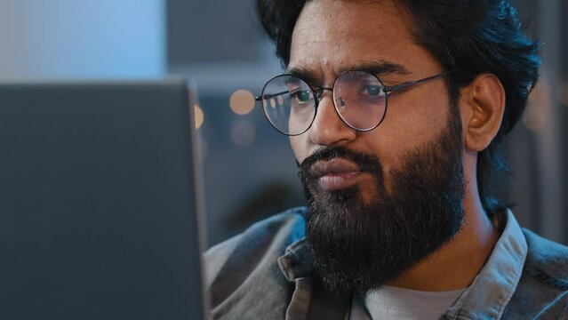 Close-up bearded male face reflection light from laptop monitor in glasses. Busy serious focused Arab Indian man looking at computer working freelance in evening playing game at night surfing in net