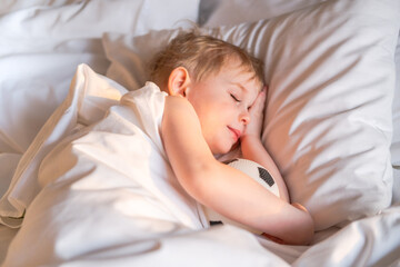 Toddler boy sleeping with a soccer ball on a white bed linen.