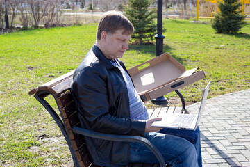 An adult blond man opens a box with a new laptop while sitting bench in the park.