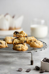 Chocolate chip cookies on a cooking lattice on a light gray concrete background, with a glass of milk, a plate of muffins and a bowl of chocolate drops