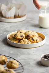 Plate with classic chocolate chip cookies on a concrete gray background with a glass of milk and muffins. Sweet crunchy biscuits for a morning snack