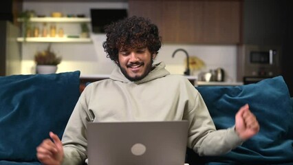 Cheerful Indian man wearing casual hoodie sitting on the sofa and using laptop at home. Inspired male entrepreneur typing on keyboard and looks aside with pleasure smile, taking a break and resting