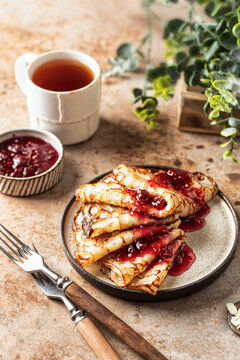 Crepes with raspberry jam topping in brown plate. Breakfast composition with forks, tea