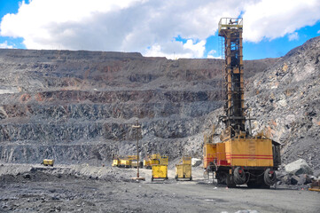The process of mining iron ore. A drilling rig is drilling holes, an excavator is loading a large dump truck.