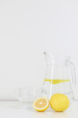 Bright image of refreshing healthy natural citrus lemon water, lemonade, in glass jug and cup besides a full and sliced lemon fruit, on white table against white wall background