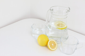 Refreshingly healthy natural citrus lemon water in a glass jug along with glass cups, and a full and sliced lemon