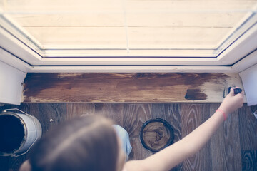 Kid girl with pigtails paints, covers a wooden board threshold with dark impregnation (oil), top view. Home renovation and creativity