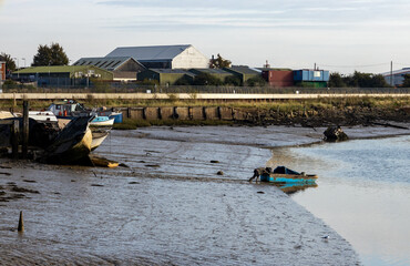 A small working boat is pushed into the River Medway in Rochester, on a sunny October morning in 2021.  A number of other boats are on the muddy foreshore, with industrial buildings in the background.