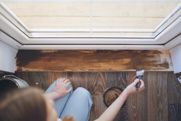 Kid girl with pigtails paints, covers a wooden board threshold with dark impregnation (oil), top...