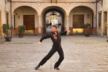 Young man with beard and ponytail, wearing black transparent shirt with black polka dots and red roses, black pants and jacket, dancing flamenco in the city. Concept art, dance, culture, tradition.
