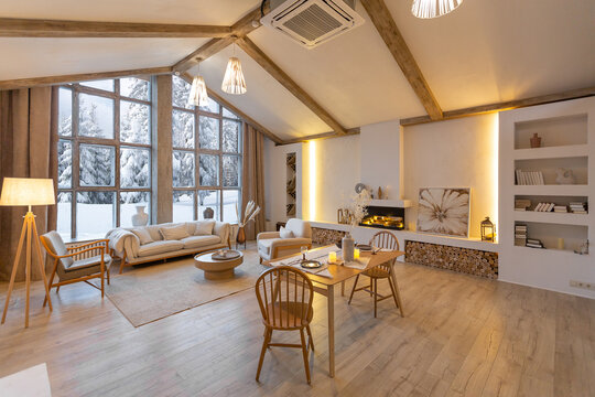 cozy warm home interior of a chic country chalet with a huge panoramic window overlooking the winter forest. open plan, wood decoration, warm colors and a family hearth