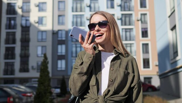 Caucasian beautiful young female blonde in sunglasses standing in urban city outdoors and recording voice message on smartphone. Stylish woman talking and sending audio record on mobile phone.