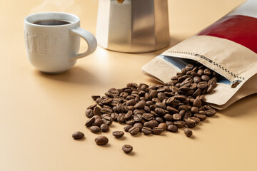 Organic coffee beans spilled out of a brown paper bag on a beige background. Whole roasted coffee beans, cup of hot coffee and a geyser coffee maker on a table. Making tasty strong coffee.