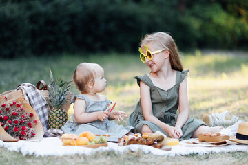 Candid lifestyle portrait of two caucasian siblings eating healthy food on picnic blanket at summer. Cute baby one year old and her sister seven years old having fun together during vacations on