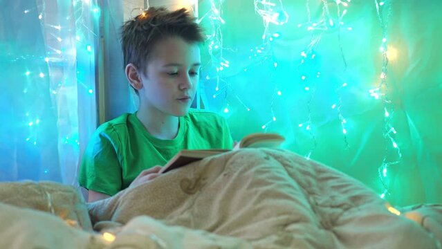 a boy is reading a book sitting on a bed decorated with garlands. multicolored light of lights. a teenager is sitting under a blanket