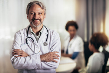 Smiling mature doctor wearing stethoscope and white lab medical coat standing in office.