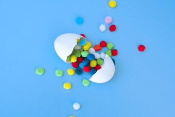 Egg with multi-colored pompoms poured out of it on a blue background, top view