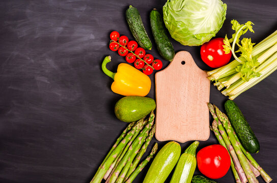 Fresh vegetables for healthy eating around a cutting board with place for text on a black background.
