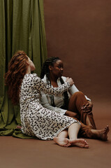 Young white woman bonding her black girlfriend while they sitting on floor against the brown background