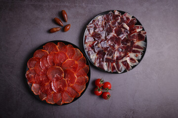 Portions of acorn-fed Iberian loin and 100% Dehesa de Extremadura acorn-fed Iberian ham on a black plate and gray table, decorated with tomatoes and acorns - obrazy, fototapety, plakaty
