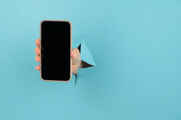 A female hand sticking out of a hole from a blue background holds a smartphone with a black screen. 