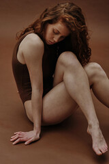 Fototapeta premium Young ginger woman with slim body sitting in swimsuit against the brown background