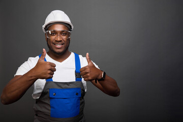 Smiling, dark-skinned worker in a gray helmet and construction jumpsuit with a blue pocket shows his thumbs up with two hands. A joyful man in overalls poses against a gray wall - obrazy, fototapety, plakaty