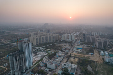 Fototapeta premium aerial drone shot passing over a building with homes, offices, shopping centers moving towards skyscapers in front of sunset showing the empty outskirts of the city of gurgaon