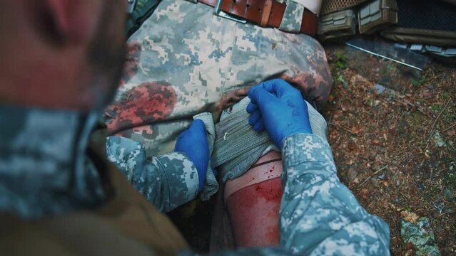 Close up hands combat medic with gloves fixes the wounded leg with bandage. Blood around. Give first aid. Military concept. Training