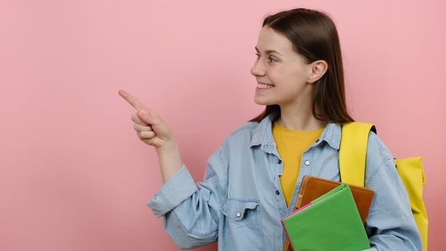 Promoter fun girl teen student 20s wears shirt and yellow backpack hold books pointing aside on workspace area copy space mock up, posing isolated over pastel pink color background wall in studio