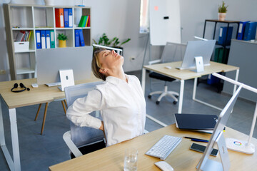 Mature business woman sits at her workplace in the office and holds on to her lower back. Health...