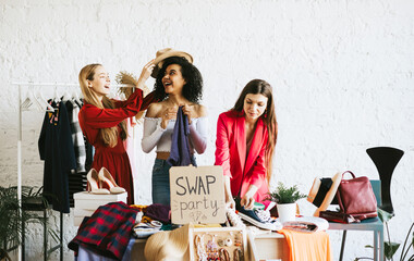 three young woman female caucasian and african students at swap party try on clothes, bags, shoes...