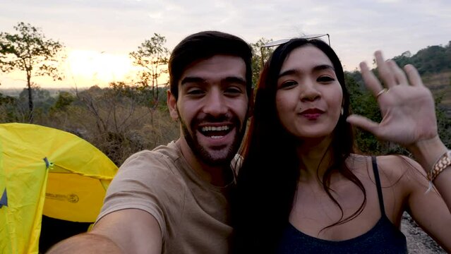 Happy love couple take photo and selfie during camping and relax in forest, beautiful asian woman and caucasian man happiness spend time tpgether in front of tent in evening.