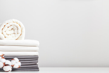 A stack of gray and white linens, sheets and a roll of towels on a table with a branch of cotton.
